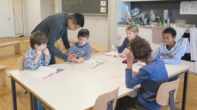 Grosser Schüler und fünf Kinder an einem Tisch in einem Schulzimmer Grosser Schüler und fünf Kinder an einem Tisch in einem Schulzimmer