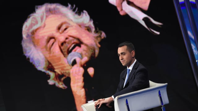 Di Maio sitzt auf einem Stuhl, hinter ihm eine grosse Projektion von Beppe Grillo. Di Maio sitzt auf einem Stuhl, hinter ihm eine grosse Projektion von Beppe Grillo.