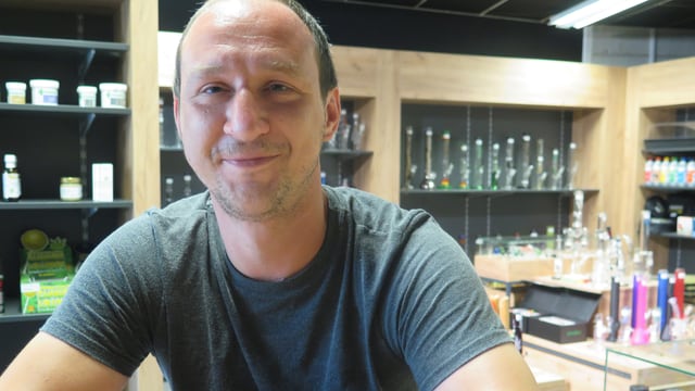 CBD-Shop-Betreiber sitzt in seinem Shop an einem Tisch. Im Hintergrund sind verschiedene Raucherprodukte zu sehen. CBD-Shop-Betreiber sitzt in seinem Shop an einem Tisch. Im Hintergrund sind verschiedene Raucherprodukte zu sehen.