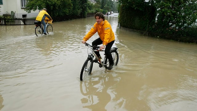 Eine überschwemmte Quartierstrasse, auf der zwei Fahrräder durch die Wassermassen pflügen. Das Wasser steht den Velofahrern dabei fast bis zu den Pedalen. Eine überschwemmte Quartierstrasse, auf der zwei Fahrräder durch die Wassermassen pflügen. Das Wasser steht den Velofahrern dabei fast bis zu den Pedalen.