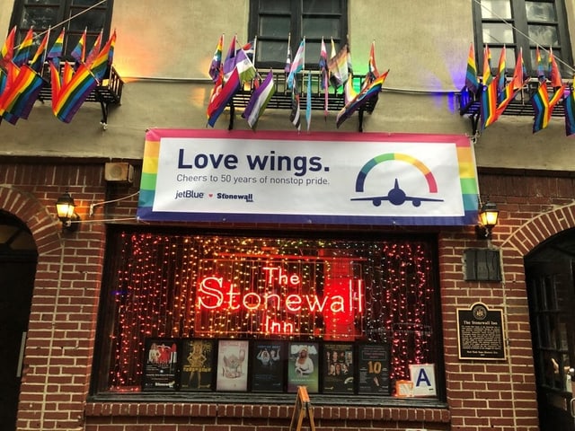Ein Bild der Stonewall Bar von aussen - über dem Fenster hängt ein Transparent auf dem steht "Love wings. Cheers to 50 years of nonstop pride. JetBlue" Ein Bild der Stonewall Bar von aussen - über dem Fenster hängt ein Transparent auf dem steht "Love wings. Cheers to 50 years of nonstop pride. JetBlue"