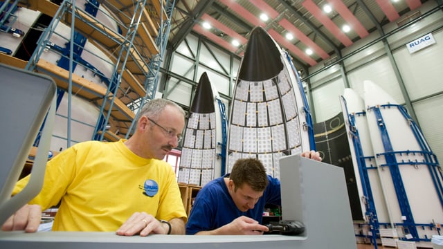 Zwei Männer arbeiten an einem Teil, im Hintergrund stehen Teile einer Raketen-Spitze. Zwei Männer arbeiten an einem Teil, im Hintergrund stehen Teile einer Raketen-Spitze.