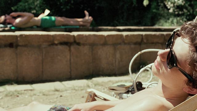 Ein Mann liegt in Badehosen in einem Liegestuhl, im Hintergrund ein anderer am Rand eines Pools. Ein Mann liegt in Badehosen in einem Liegestuhl, im Hintergrund ein anderer am Rand eines Pools.