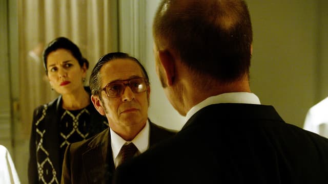 Mann mit grosser Brille (frontal) schaut einem andern Mann ins Gesicht (von hinten sichtbar). Hinten im Bild steht zudem eine Frau. Mann mit grosser Brille (frontal) schaut einem andern Mann ins Gesicht (von hinten sichtbar). Hinten im Bild steht zudem eine Frau.