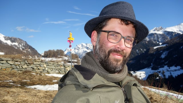 Ein Mann mit Hut und Brille vor einer Bergkulisse.