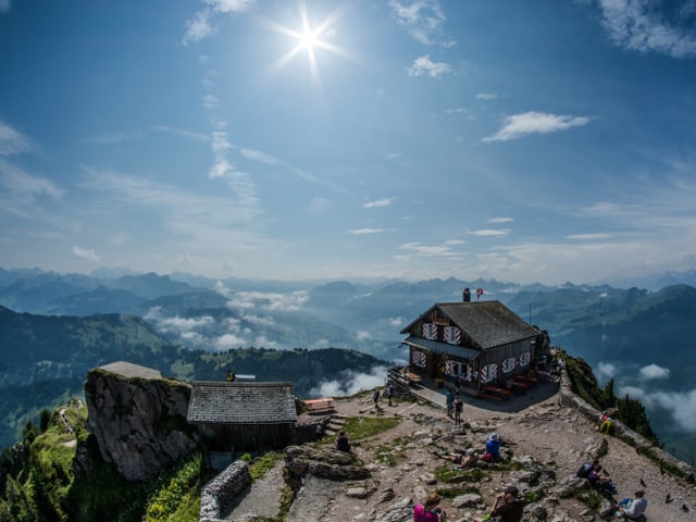 Blick auf einen Berggipfel bei strahlendem Sonnenschein. Auf dem Gipfel ist eine kleine Hütte zu sehen mit einigen Wanderen, die die Aussicht geniessen.