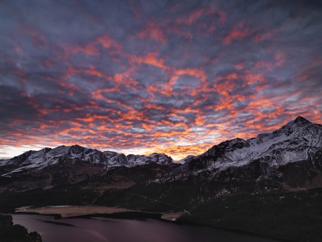 Rote bis rosa Wolken am Himmel über einer schroffen Bergwelt mit wenig Schnee auf den Gipfeln.