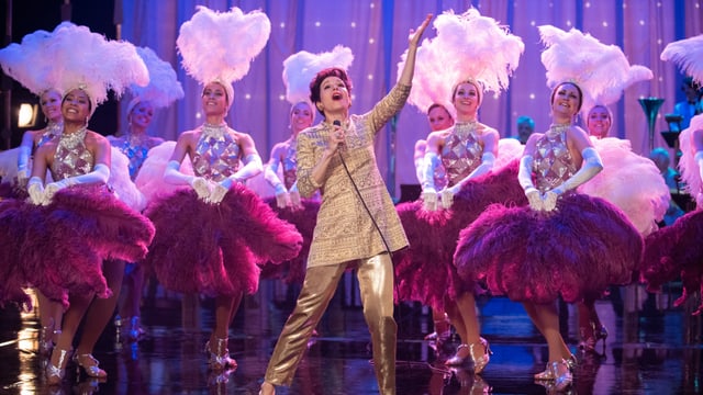 Judy steht in einem goldenen Anzug mit vielen pink angezogenen Tänzerinnen auf der Bühne. Judy steht in einem goldenen Anzug mit vielen pink angezogenen Tänzerinnen auf der Bühne.