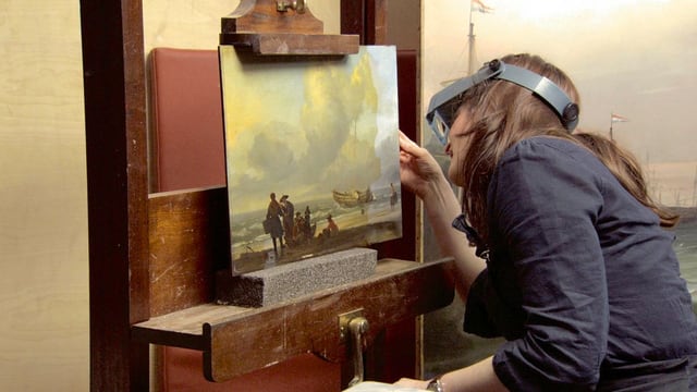 Eine Restauratorin beugt sich über ein Gemälde. Sie trägt eine spezielle Brille. Eine Restauratorin beugt sich über ein Gemälde. Sie trägt eine spezielle Brille.