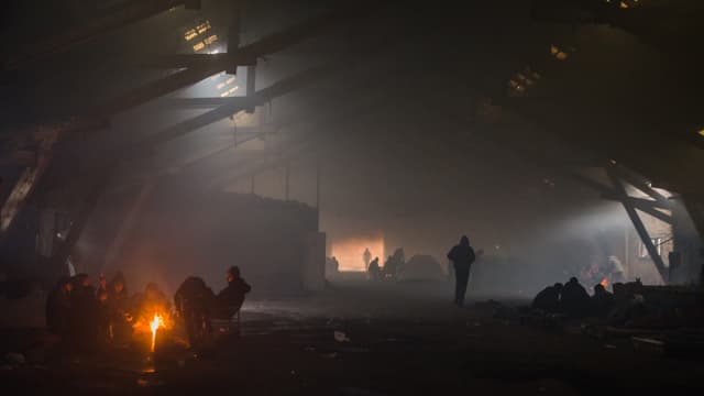 Blick in eine dunkle Lagerhalle, vorne links brennt ein Feuer. Männer sitzen darum herum. Blick in eine dunkle Lagerhalle, vorne links brennt ein Feuer. Männer sitzen darum herum.