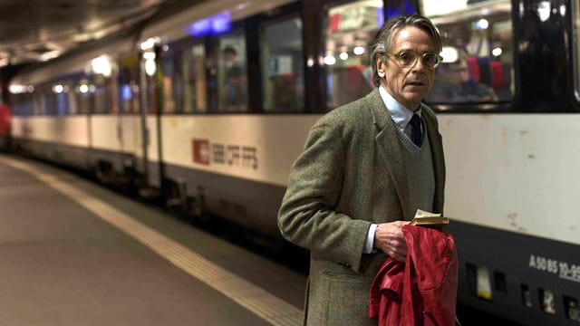 Der Schauspieler Jeremy Irons steht in einem grau-brauenen Anzug vor einem SBB-Zug.