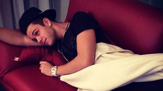 Luca Hänni seitlich und mit einer Decke zugedeckt auf einer roten Couch liegend.