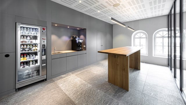 Purtret d'in local cun en ina maisa auta, ina cuschina cun maschina da cafe ed in automat cun bavrondas e snacks. Purtret d'in local cun en ina maisa auta, ina cuschina cun maschina da cafe ed in automat cun bavrondas e snacks.