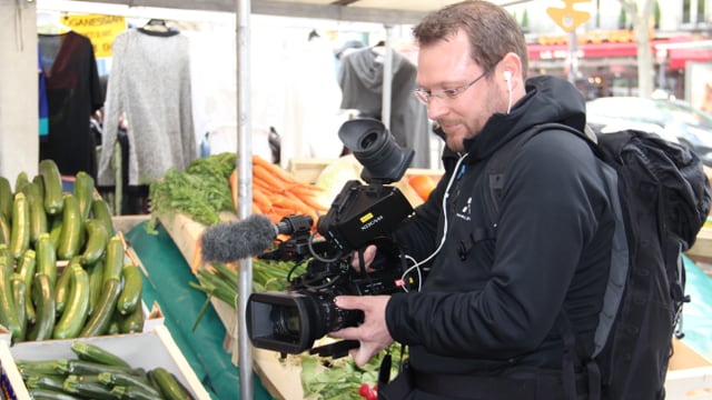 Purtret da Stefan Dobler. El filmescha cun ina camera da VJ ina fiera. Davostiers in stand cun legums. Purtret da Stefan Dobler. El filmescha cun ina camera da VJ ina fiera. Davostiers in stand cun legums.