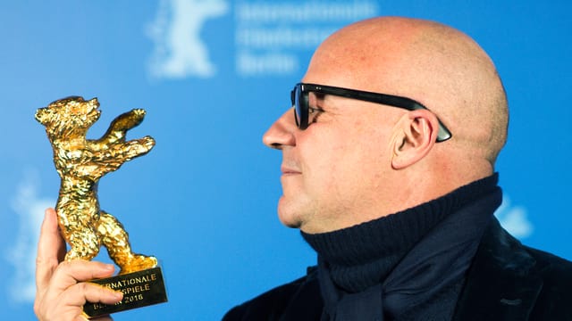 Ein Mann mit Glatze und schwarzer Brille hält die Statue eines goldenen Bären in der Hand. Ein Mann mit Glatze und schwarzer Brille hält die Statue eines goldenen Bären in der Hand.