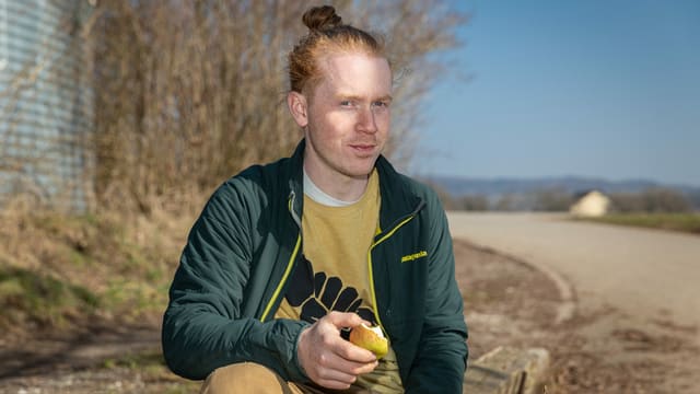 ein junger Mann sitzt draussen, isst einen Apfel und schaut in die Kamera ein junger Mann sitzt draussen, isst einen Apfel und schaut in die Kamera