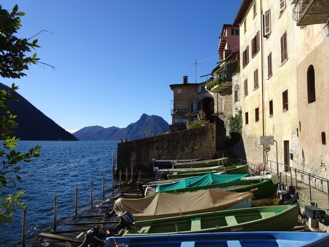 Am Ufer des Lago di Lugano mit einem Bootssteg. Der Himmel im Hintergrund erstrahlt im tiefsten Blau.