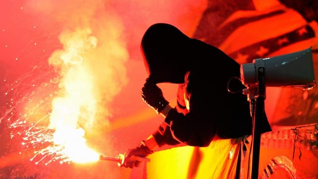 Ein Hooligan zündet eine rot leuchtende Petarde, sein Gesicht ist vermummt. Ein Hooligan zündet eine rot leuchtende Petarde, sein Gesicht ist vermummt.