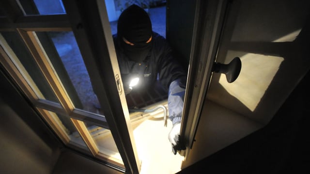 Ein Einbracher mit Maske und Taschenlampe öffnet von aussen ein Fenster Ein Einbracher mit Maske und Taschenlampe öffnet von aussen ein Fenster