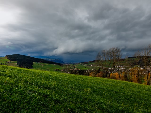 Dunkle Wolken ziehen über der hügeligen Landschaft auf. Im Vordergrund sieht man eine grüne Wiese.