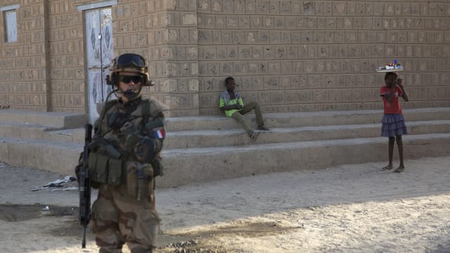 Ein Soldat in voller Kampfmontur mit gesenktem Maschinengewehr im Vordergrund. Dahinter zwei Kinder vor einer Hausmauer. Ein Soldat in voller Kampfmontur mit gesenktem Maschinengewehr im Vordergrund. Dahinter zwei Kinder vor einer Hausmauer.