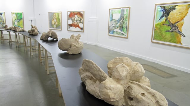 Sieben Holzskulpturen in der Mitte des Raumes, an der Wand hängen Pastell-Bilder. Sieben Holzskulpturen in der Mitte des Raumes, an der Wand hängen Pastell-Bilder.
