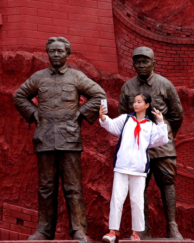 Eine junge Chinesin steht vor einer Mao-Statue und einem Relief der chinesischen Mauer und macht ein Selfie.
