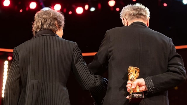 Eine Frau und ein Mann stehen mit dem Rücken zur Kamera, er hält einen goldenen Bären Eine Frau und ein Mann stehen mit dem Rücken zur Kamera, er hält einen goldenen Bären