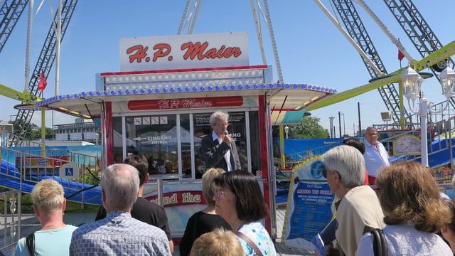 Ein Mann steht vor einem Riesenrad und redet in ein Mikrofon. Ein Mann steht vor einem Riesenrad und redet in ein Mikrofon.
