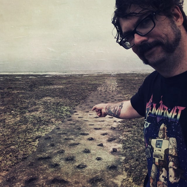 Guido steht am Strand und zeigt auf den Kabelkanal aus Beton. Er trägt ein T-Shirt mit der Aufschrift «Internet». Guido steht am Strand und zeigt auf den Kabelkanal aus Beton. Er trägt ein T-Shirt mit der Aufschrift «Internet».