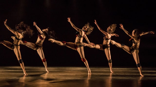 Ausschnitt aus einer Gruppenchoreographie, in der  die exotische Volkstümlichkeit durchwegs zeitgenössisch interpretiert wird: fünf Tänzerinnen auf der Bühne.