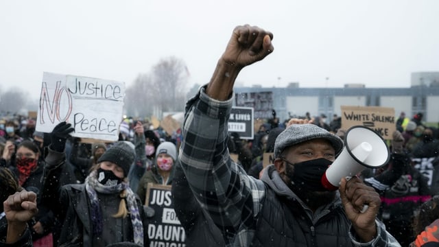 Viele Menschen mit Maske halten Schilder oder ihre Fäuste in die Höhe. Ein Mann spricht in ein Megafon. Viele Menschen mit Maske halten Schilder oder ihre Fäuste in die Höhe. Ein Mann spricht in ein Megafon.