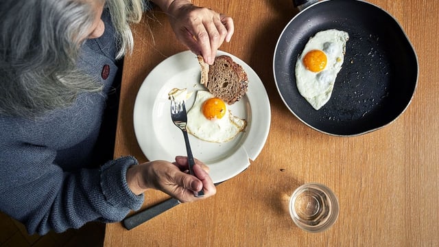 Eine ältere Frau sitzt am Tisch und isst Spiegeleier mit Brot. Eine ältere Frau sitzt am Tisch und isst Spiegeleier mit Brot.