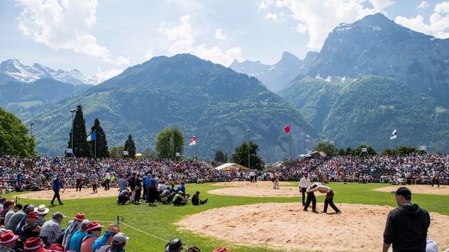 Im Vordergrund die Schwingplätze aus Sägemehl auf einer Wiese. Rundherum Zuschauer auf Tribühne. Im Hintergrund Bergpanorama. Im Vordergrund die Schwingplätze aus Sägemehl auf einer Wiese. Rundherum Zuschauer auf Tribühne. Im Hintergrund Bergpanorama.