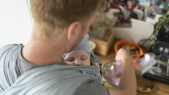 Vater bereitet Essen zu, während Baby in Tragetuch an seiner Brust schläft