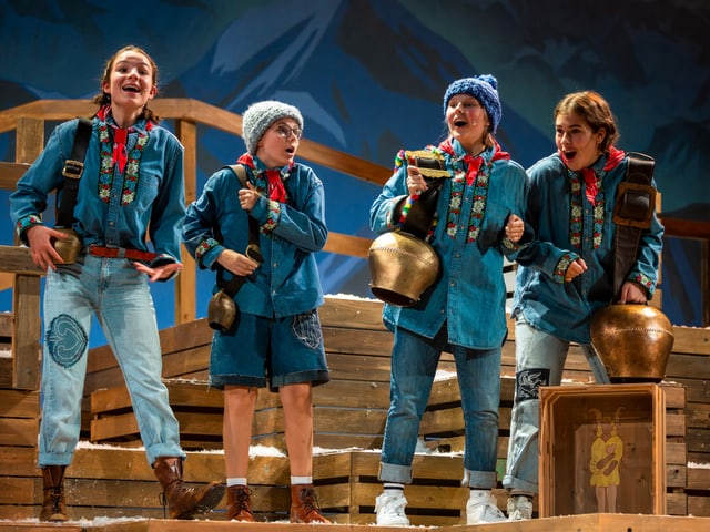 Vier Kinder in traditioneller Schweizer Kleidung stehen auf Holzkisten und singen. Alle tragen eine Glocke um den Arm. Sie spielen Kinder aus dem Kinderbuchklassiker Schellen-Ursli Vier Kinder in traditioneller Schweizer Kleidung stehen auf Holzkisten und singen. Alle tragen eine Glocke um den Arm. Sie spielen Kinder aus dem Kinderbuchklassiker Schellen-Ursli