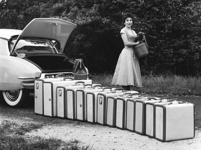 Eine Frau mit sehr vielen Koffern steht hinter einem Auto.