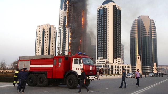 Ein Hochhaus steht in Flammen. Im Bildvordergrund ist ein rotes Feuerwehrauto zu sehen. Ein Hochhaus steht in Flammen. Im Bildvordergrund ist ein rotes Feuerwehrauto zu sehen.