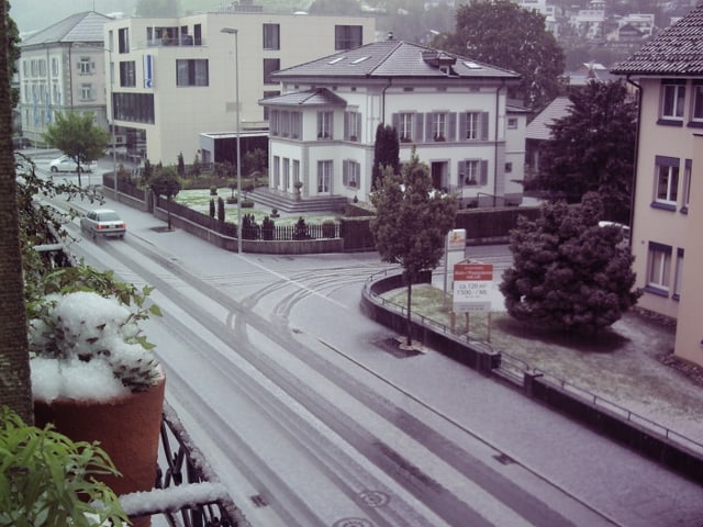Blick vom ersten Stock auf die Strasse in einem Dorf. Alles ist weiss. Es hat überall Graupelkörner. Die Strassen scheinen glatt zu sein.