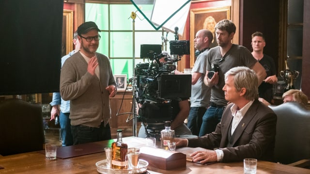 Filmset: Ein Mann sitzt am Tisch, ein Mann steht links von ihm, hat die Hand erhoben und scheint etwas zu erklären. Um die beiden herum stehen die Kameras und ein Film-Team. Filmset: Ein Mann sitzt am Tisch, ein Mann steht links von ihm, hat die Hand erhoben und scheint etwas zu erklären. Um die beiden herum stehen die Kameras und ein Film-Team.