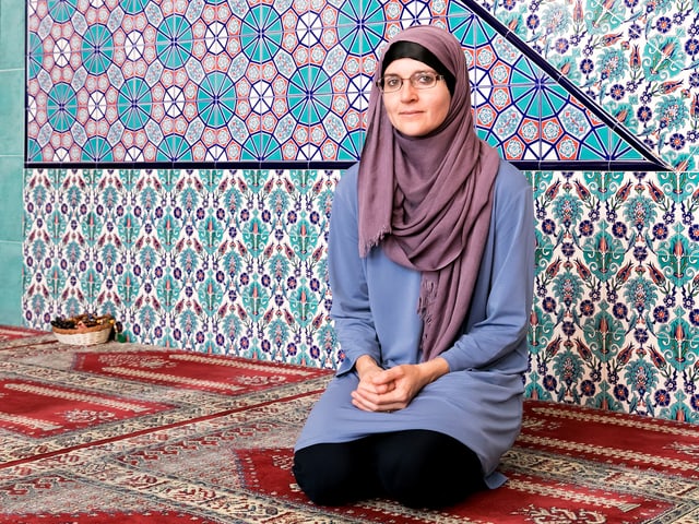 Eine Frau mit glaubem Kleid und Kopftuch sitzt auf dem Teppichboden einer Moschee.