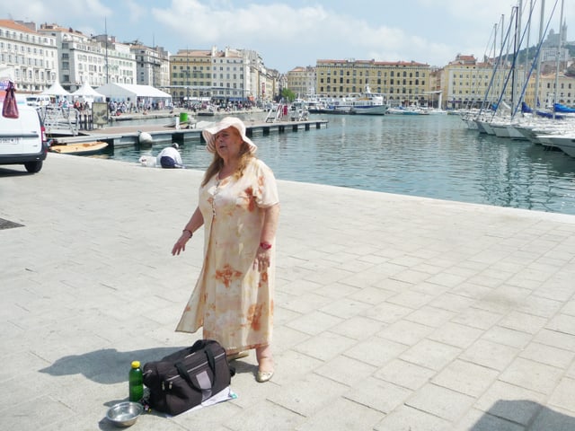 Eine Frau steht an einem Hafen und singt. Im Hintergrund sieht man Boote und Häuser von Marseille. Eine Frau steht an einem Hafen und singt. Im Hintergrund sieht man Boote und Häuser von Marseille.