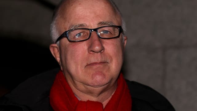Porträtaufnahme von MacShane mit rotem Schal und schwarzer Brille. Porträtaufnahme von MacShane mit rotem Schal und schwarzer Brille.