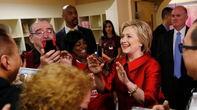 Hillary Clinton cun in deux pieces cotschen e fatscha rienta. Ella tegn ils mauns sin altezia da la schugliala. Ella è circumdada da fans e reporters. Hillary Clinton cun in deux pieces cotschen e fatscha rienta. Ella tegn ils mauns sin altezia da la schugliala. Ella è circumdada da fans e reporters.
