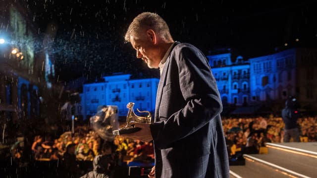 Mann mit kleiner goldener Leopardenstatuette vor Publikum. Im Scheinwerferlicht ist Regen zu erkennen. Mann mit kleiner goldener Leopardenstatuette vor Publikum. Im Scheinwerferlicht ist Regen zu erkennen.