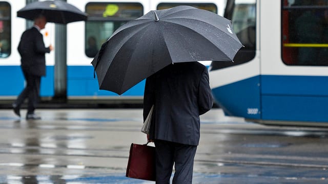 Bankmitarbeiter unter Regenschirm stehend mit einer Aktentasche in der Hand. Tram im Vordergrund Bankmitarbeiter unter Regenschirm stehend mit einer Aktentasche in der Hand. Tram im Vordergrund