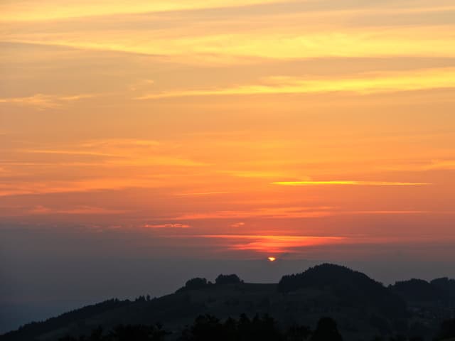 Im Vordergrund Hügel, Ausblick ins Diffuse. Der Himmel ist unten grau, dann lachsrot bis orange und oben gelb.