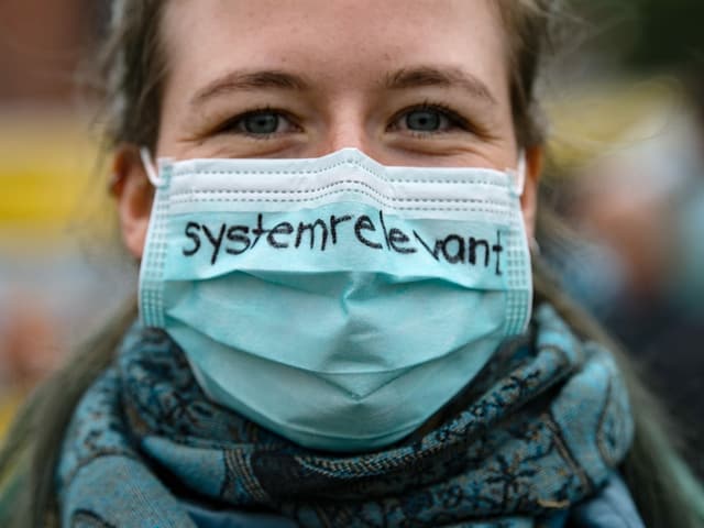 Eine Frau trägt eine türkisfarbene Mase, auf der «systemrelevant» steht. Eine Frau trägt eine türkisfarbene Mase, auf der «systemrelevant» steht.