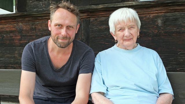 Mann neben alter Frau auf Bank vor Holzhaus. Mann neben alter Frau auf Bank vor Holzhaus.