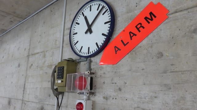Eine Uhr an der Wand, darunter ein Telefon und ein roter Knopf, auf den ein grosses Schild weist. Auf diesem steht: Alarm. Eine Uhr an der Wand, darunter ein Telefon und ein roter Knopf, auf den ein grosses Schild weist. Auf diesem steht: Alarm.